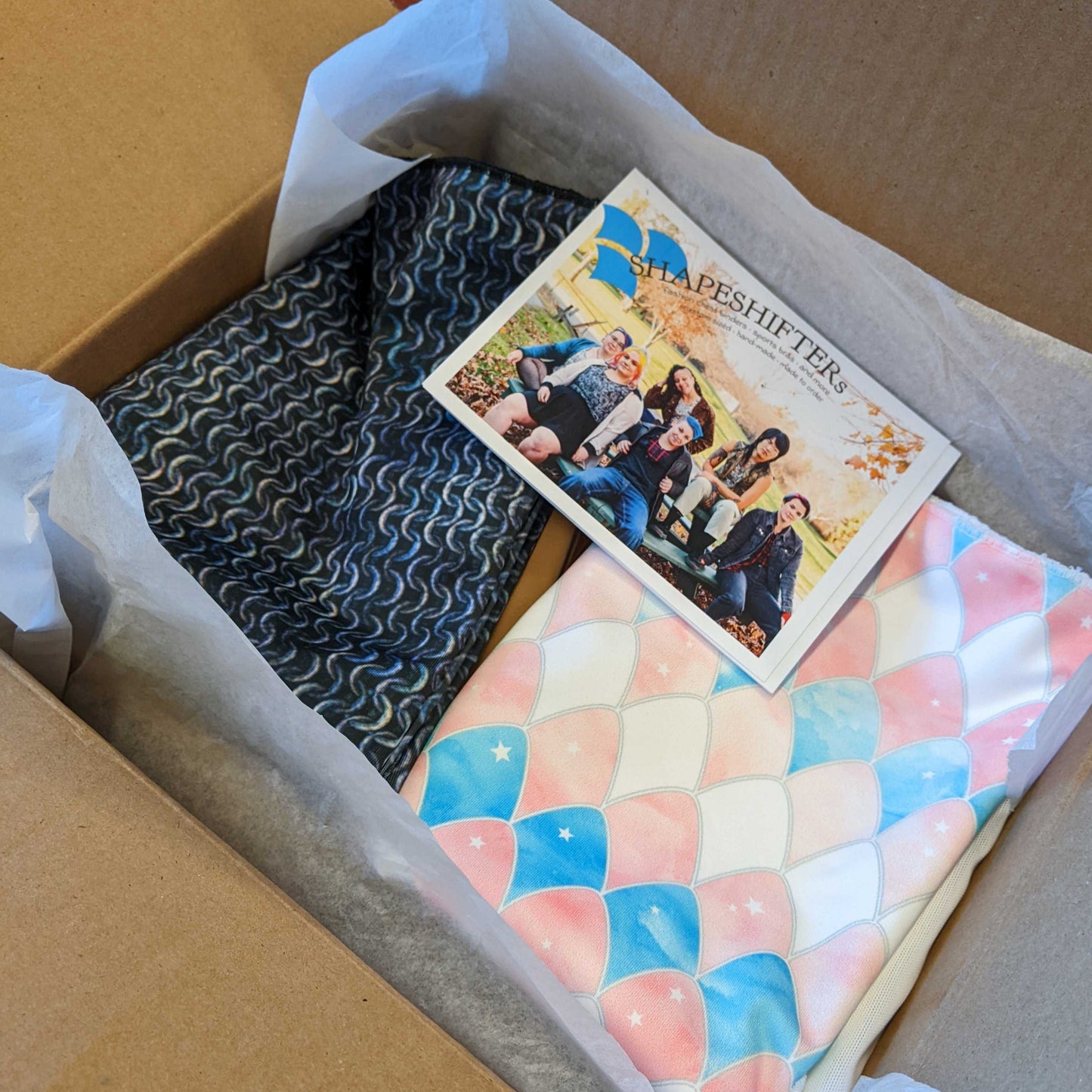 A box of packed binders, with a chainmail print and a trans dragon scale print on top.